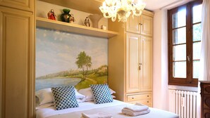 A bedroom with a bed, pillows, a painting of a landscape, a chandelier, and a window. - Wine Apartments Florence Soave (Firenze)