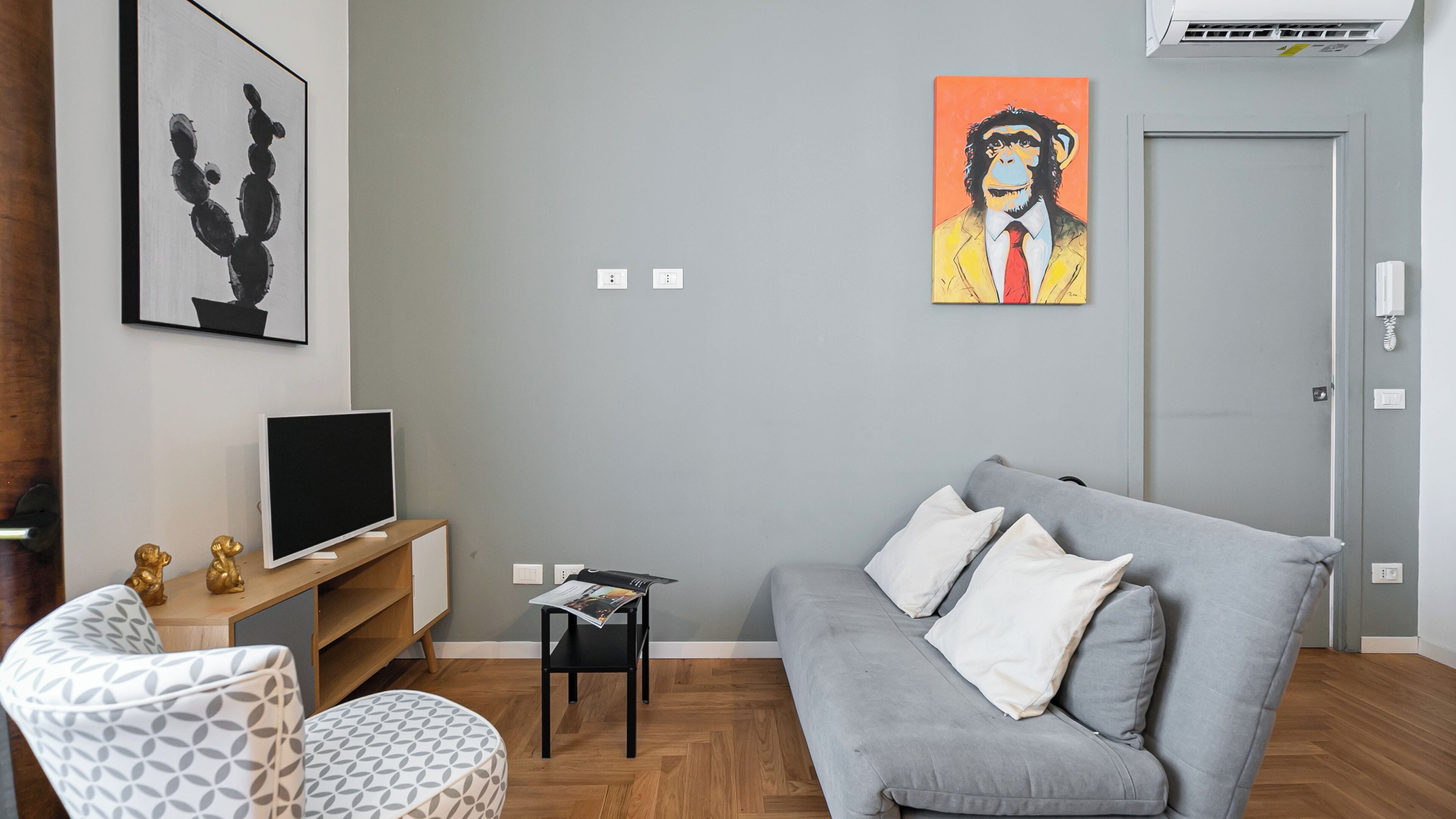 Un salon moderne avec un canapé gris, un téléviseur posé sur un meuble en bois, une petite table avec des magazines et des œuvres d’art accrochées au mur.