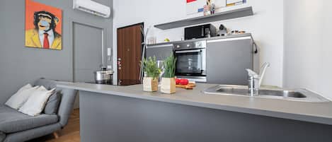 A modern kitchen with a grey countertop, stainless steel appliances, and a grey sofa in the living area.