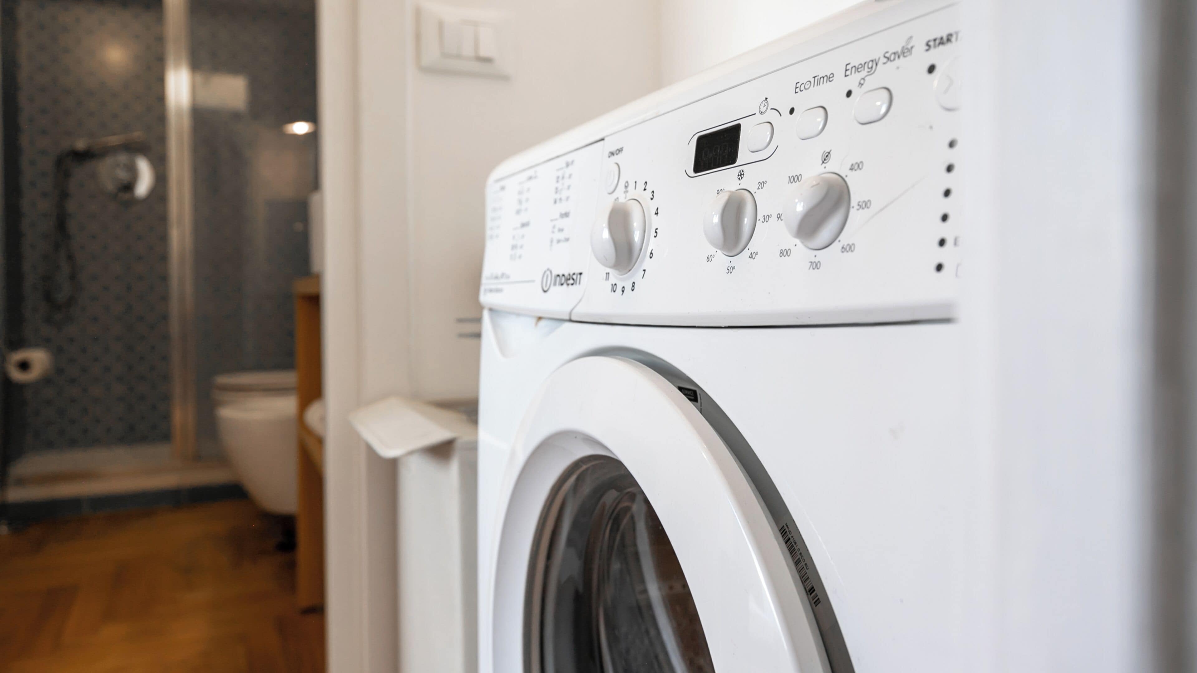 A front view of a washing machine with control knobs and a digital display, located in a bathroom with a shower and toilet visible in the background.