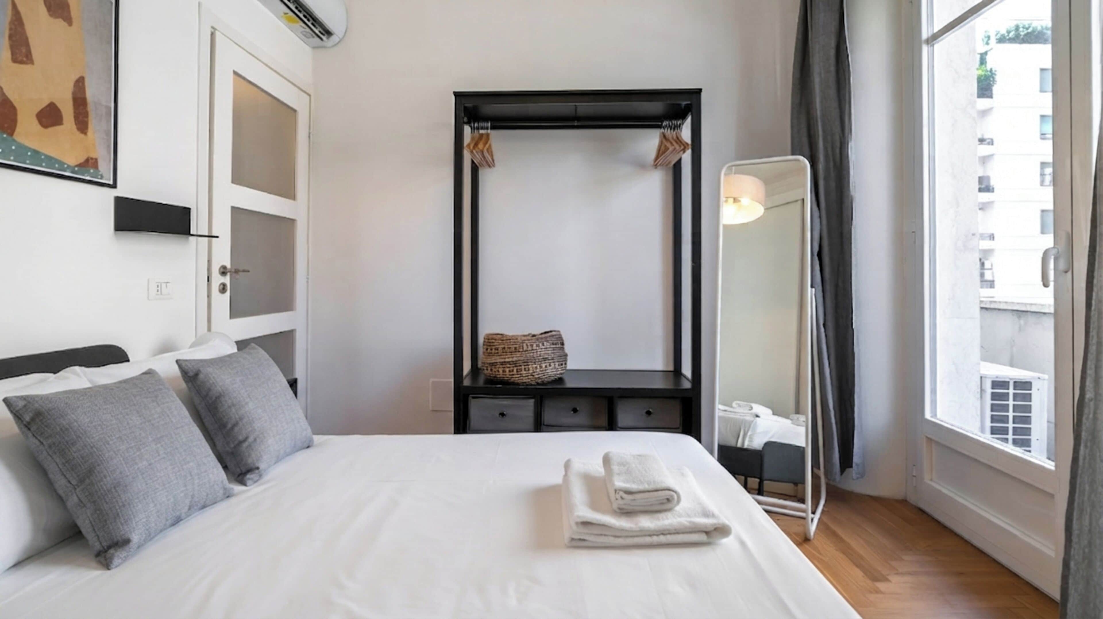Hotellihuone, jossa on sänky, yöpöytä lampun kanssa, suuri peili ja näkymä ulos.