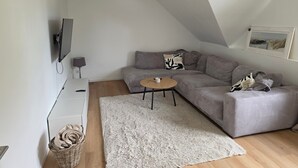 Een moderne woonkamer met een grijze bank, een klein rond salontafeltje, een wit vloerkleed en een flatscreen televisie die aan de muur is bevestigd.