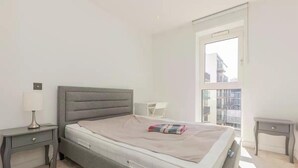Un dormitorio con cama, mesita de noche, lámpara y ventana con vista a edificios.