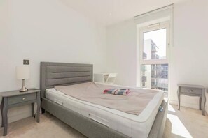 Ein Schlafzimmer mit einem Bett, einem Nachttisch, einer Lampe und einem Fenster mit Blick auf Gebäude.