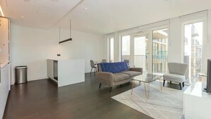 Una sala de estar moderna con un sofá, una mesa de centro y un sillón. Cuenta con una pequeña área de cocina con un refrigerador y un bote de basura. La habitación tiene ventanas amplias y un balcón.