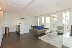 Ein modernes Wohnzimmer mit einer Couch, einem Couchtisch und einem Sessel. Es gibt eine kleine Küchenzeile mit einem Kühlschrank und einer Mülltonne. Der Raum verfügt über große Fenster und einen Balkon.