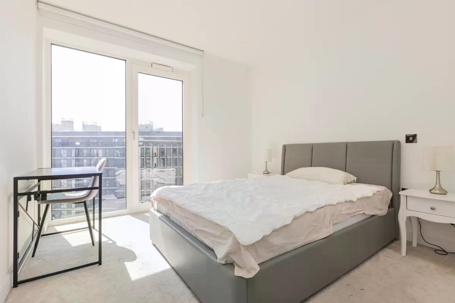 Un dormitorio con cama, mesita de noche, lámpara, silla y ventana con vista a edificios.