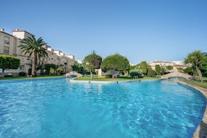 Piscina com água azul cristalina, cercada por vegetação bem cuidada e edifícios com estilo arquitetônico mediterrâneo.