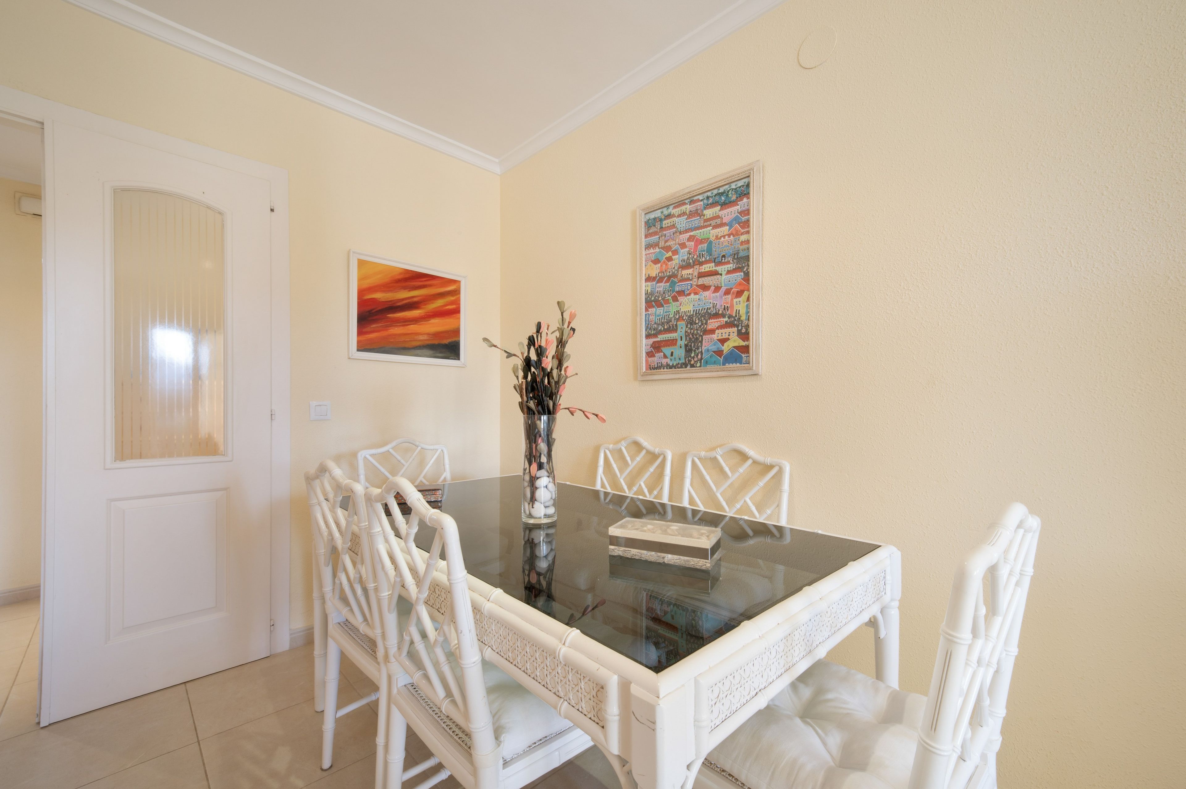 Un comedor con una mesa de vidrio, sillas blancas y dos obras de arte enmarcadas en la pared.