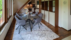 Un coin repas avec une table dressée pour quatre personnes, une cuisine en arrière-plan et une vue sur l’extérieur grâce à de grandes fenêtres.