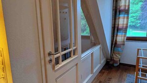 Une pièce avec un plancher en bois, une porte blanche dotée de panneaux de verre, une fenêtre avec des rideaux et une vue sur la verdure à l’extérieur.