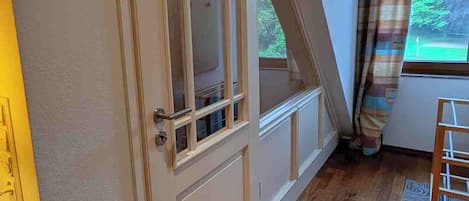 Een kamer met een houten vloer, een witte deur met glaspanelen, een raam met gordijnen en uitzicht op groen.