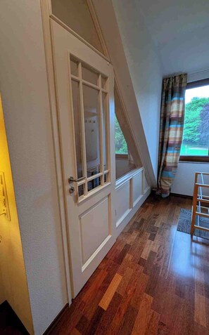 Ein Zimmer mit Holzboden, einer weißen Tür mit Glasfeldern, einem Fenster mit Vorhängen und Blick auf Grünfläche.