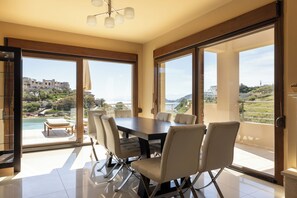 Comedor moderno con una mesa grande y sillas, ventanas amplias y vistas a una piscina y edificios.