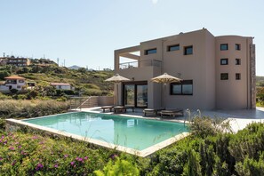 A modern building with a pool, outdoor seating, and a view of a hilly landscape. - Villa Mononaftis (Heraklion)
