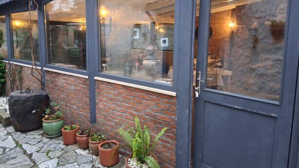 A building with a glass facade, a brick wall, and a door. There are potted plants outside. - ADATEPE OTEL KAZDAĞLARI (Ayvacik)