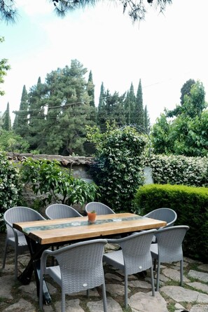 Un área de comedor al aire libre con una mesa de madera y sillas de mimbre, rodeada de follaje y árboles.