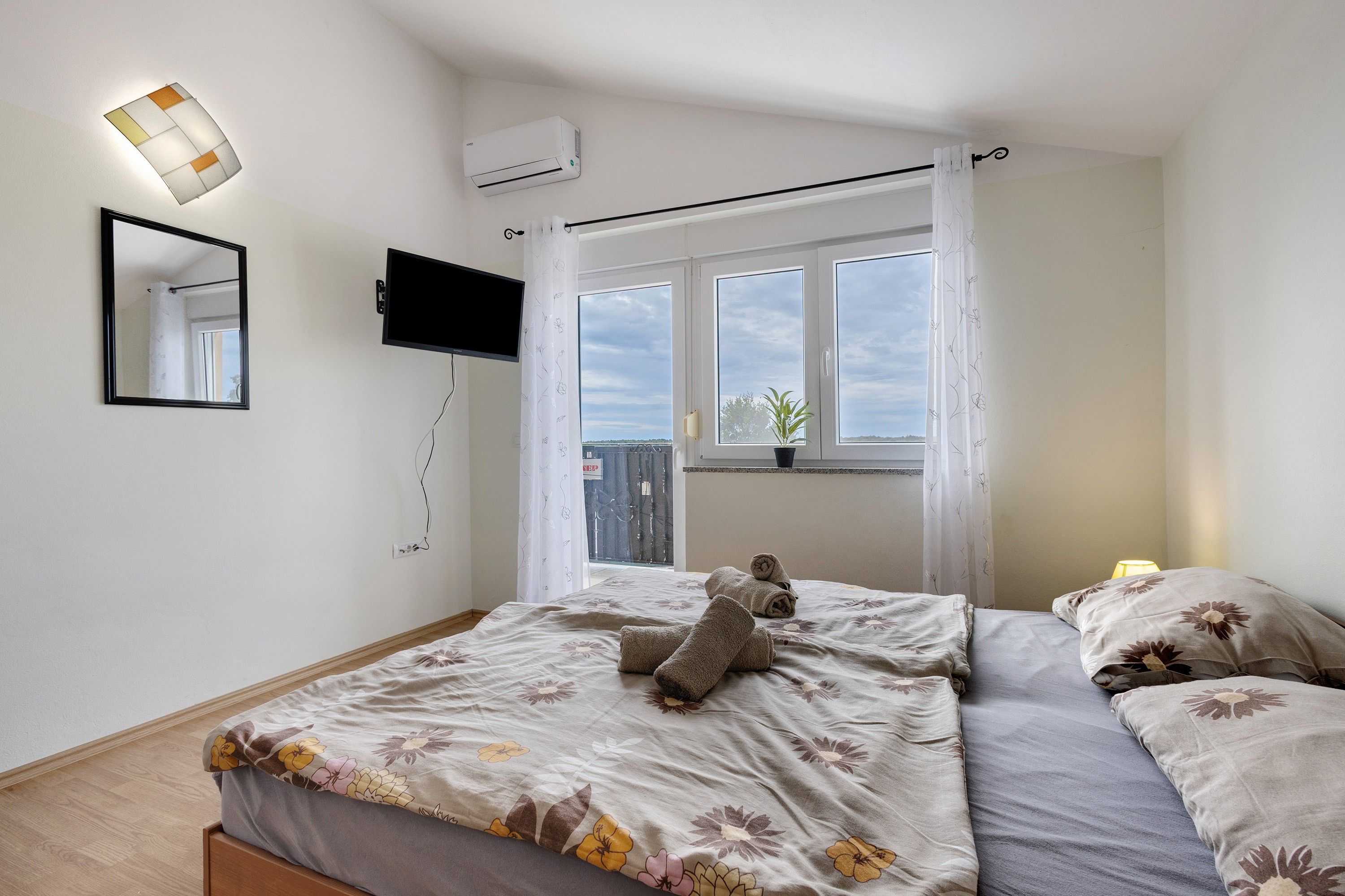 A bedroom with a bed, a flat-screen TV, a window with curtains, and an air conditioning unit.