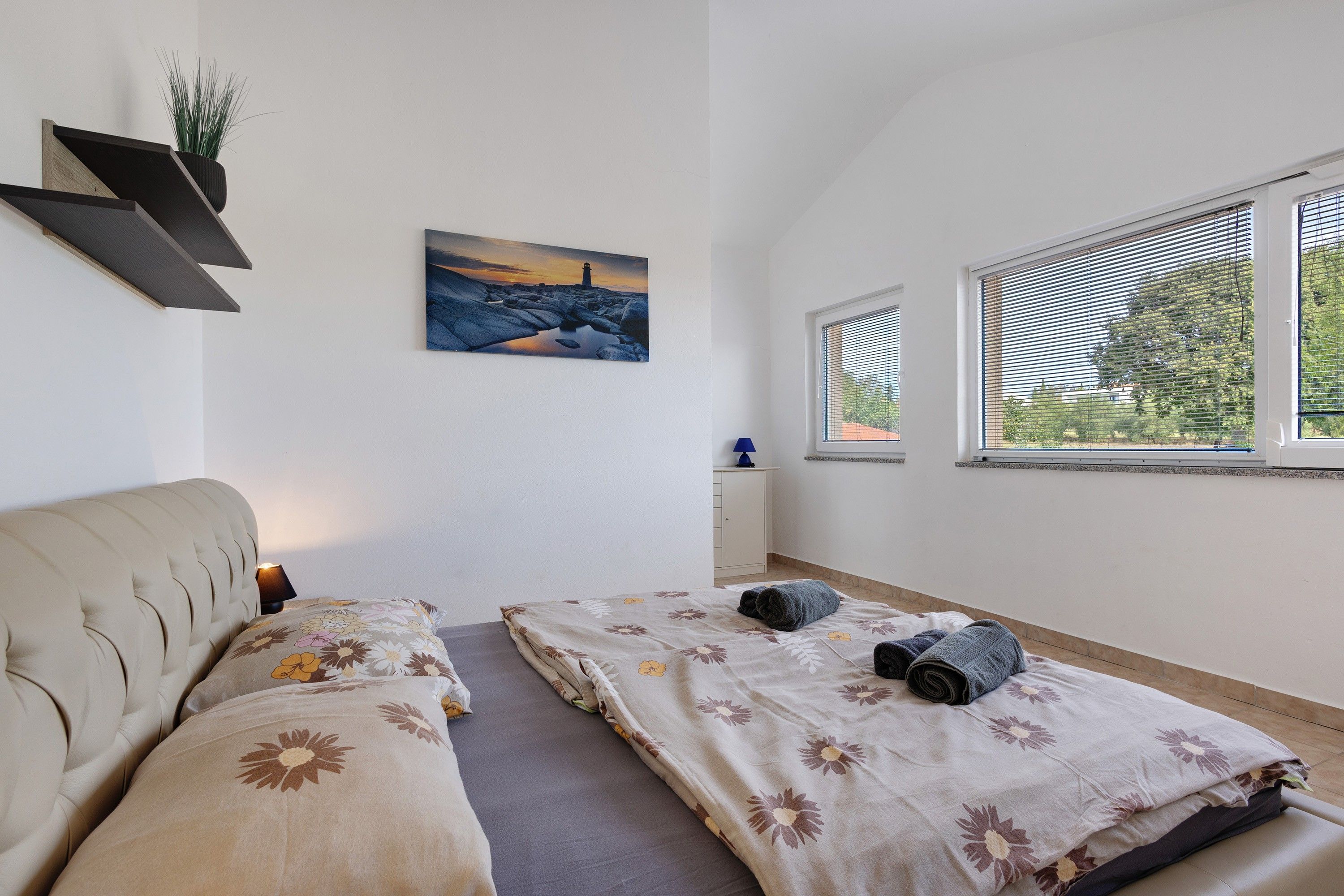 A bedroom with a bed, a sofa, a painting, a window, and a plant.