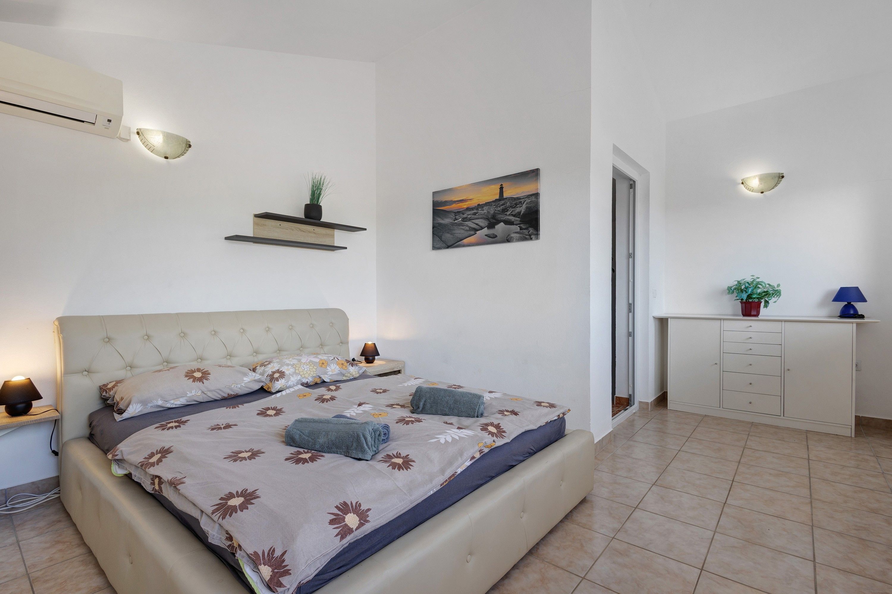 A bedroom with a bed, bedside tables, a nightstand, a wall-mounted shelf with a plant, a painting, and a cabinet.
