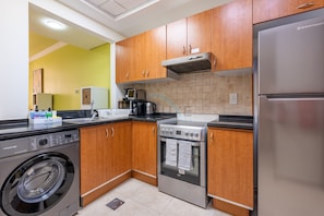 A compact kitchen with wooden cabinets, a stainless steel refrigerator, a built-in oven, a washing machine, and a coffee maker. - Canalfront Studio Steps from the Waterfront Promenade and Cricket Stadium (Dubai)