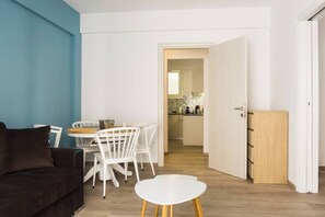 Ein modernes Wohnzimmer mit einer Couch, einem Esstisch mit Stühlen, einem kleinen weißen Tisch und einer durchgehenden Tür erkennbaren Küche.