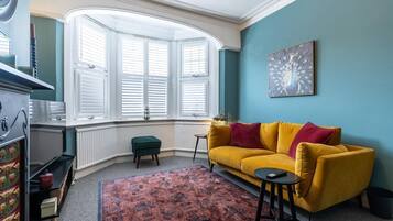 Une pièce avec une baie vitrée, un canapé jaune, un tapis à motifs et un tableau accroché au mur.