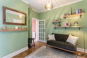 Una habitación con pared verde, piso de madera, sofá con cojines estampados, un espejo y estantes con objetos decorativos.