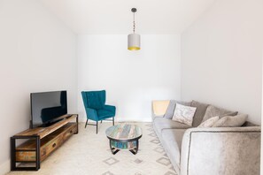 Un salon avec un téléviseur posé sur un meuble en bois, un fauteuil turquoise, une table basse ronde et un canapé gris agrémenté de coussins.