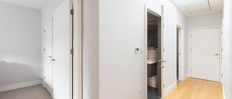 A hallway with white walls and wooden flooring, leading to a bathroom with a shower and a toilet.
