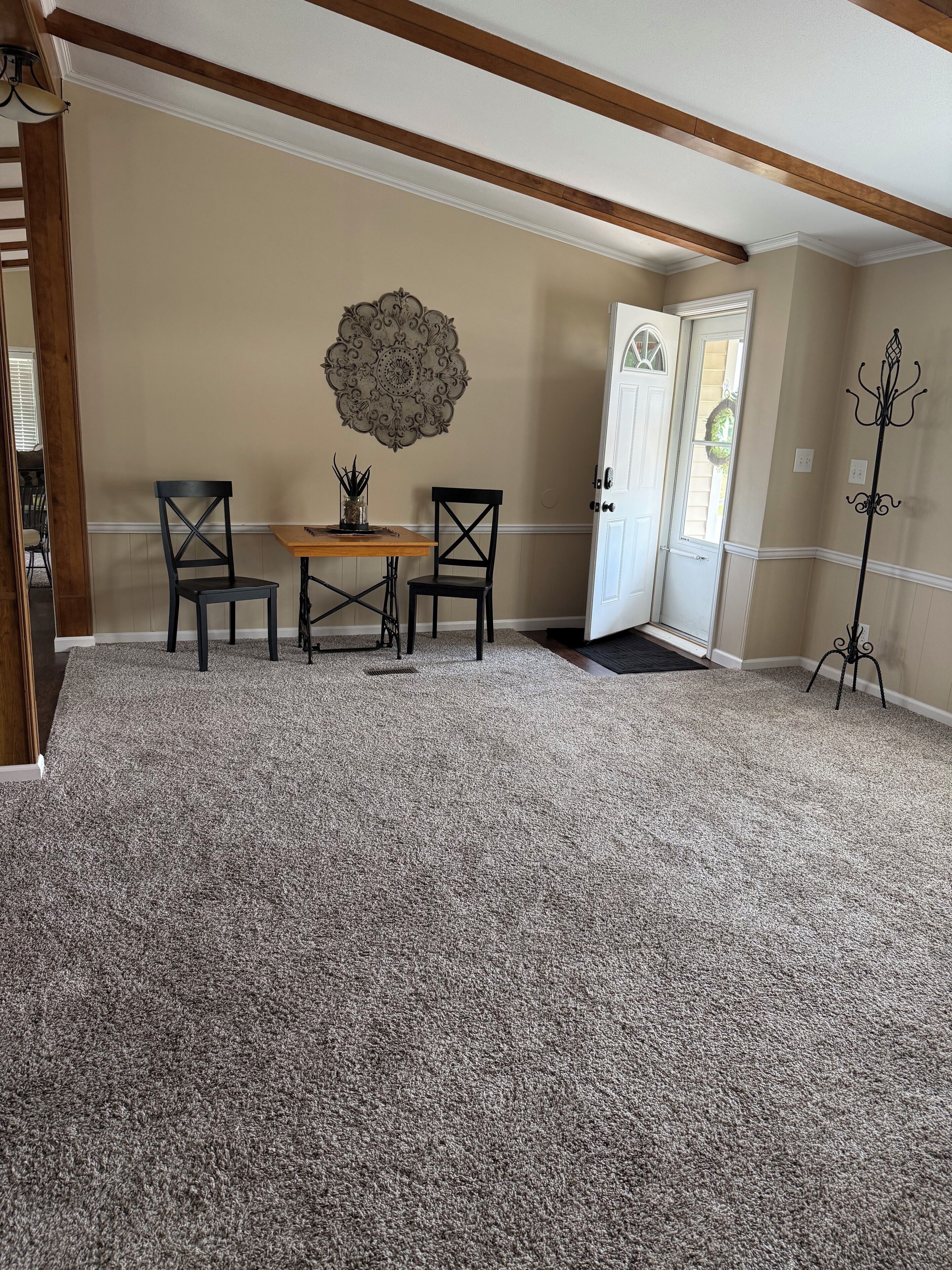 A room with a carpeted floor, a wooden table with chairs, a decorative wall piece, and a coat rack.