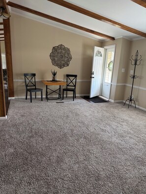 A room with a carpeted floor, a wooden table with chairs, a decorative wall piece, and a coat rack.