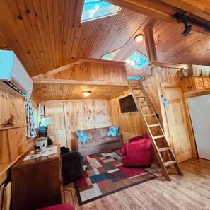 Interior acogedor de una cabaña con una cama en el entrepiso, un sofá, un escritorio y una escalera que conduce a la zona de descanso en altura.