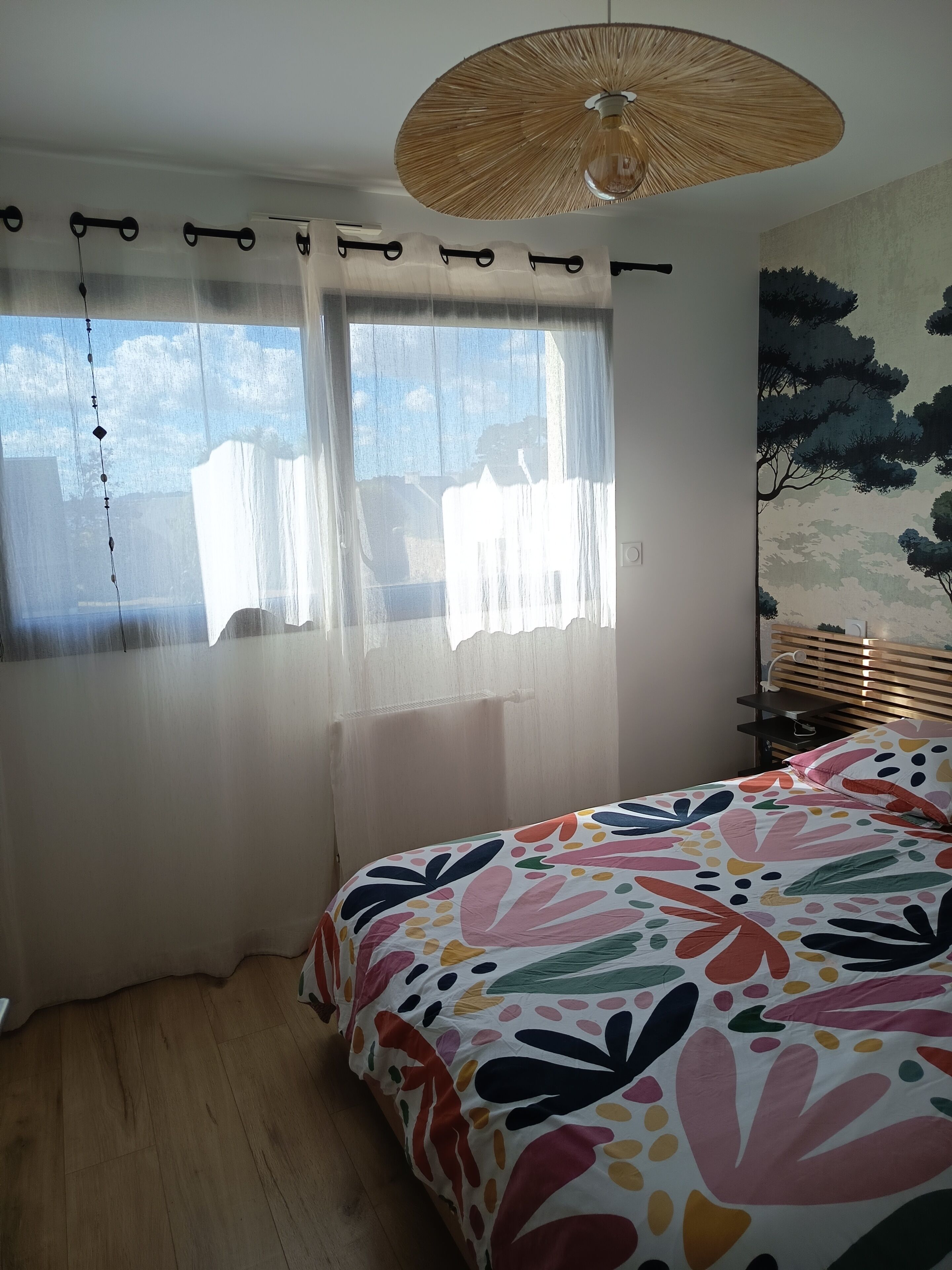 A bedroom with a bed, a window with sheer curtains, a hanging light fixture, and a wall with a tree mural.