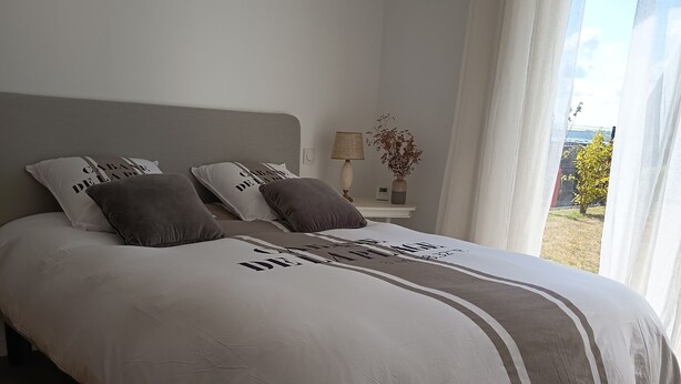 A bedroom with a large bed, a bedside table with a lamp, and a view of the outdoors. - Sea view house 5 minutes walk from the beach and coastal paths. (Saint-Pol-de-Léon)