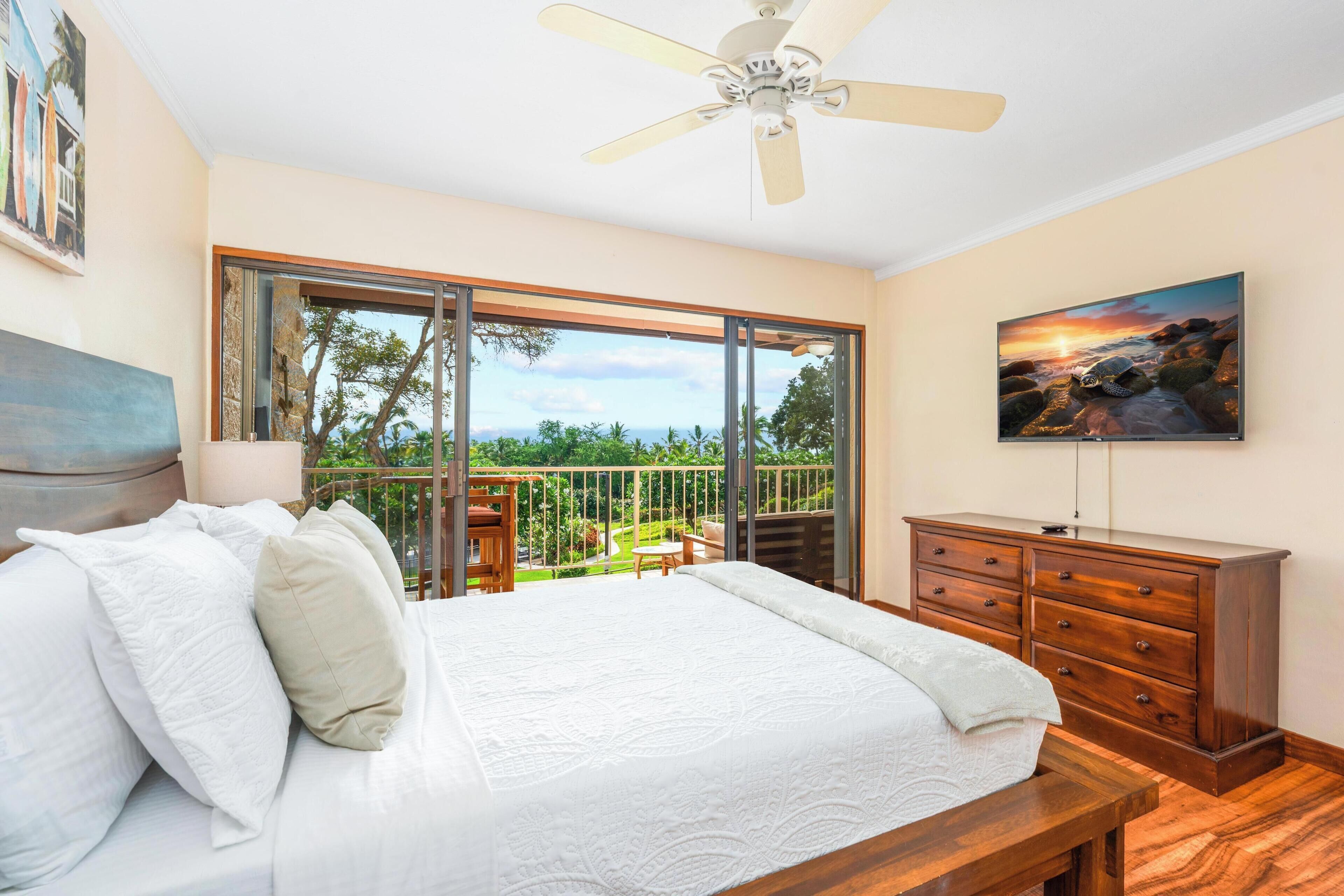 Ein Schlafzimmer mit Bett, Kommode, Deckenventilator, Fernseher und einem Balkon mit Ausblick ins Freie.