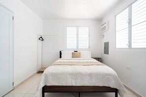 Un dormitorio con cama, mesita de noche, lámpara de pie, ventana con persianas y puerta.