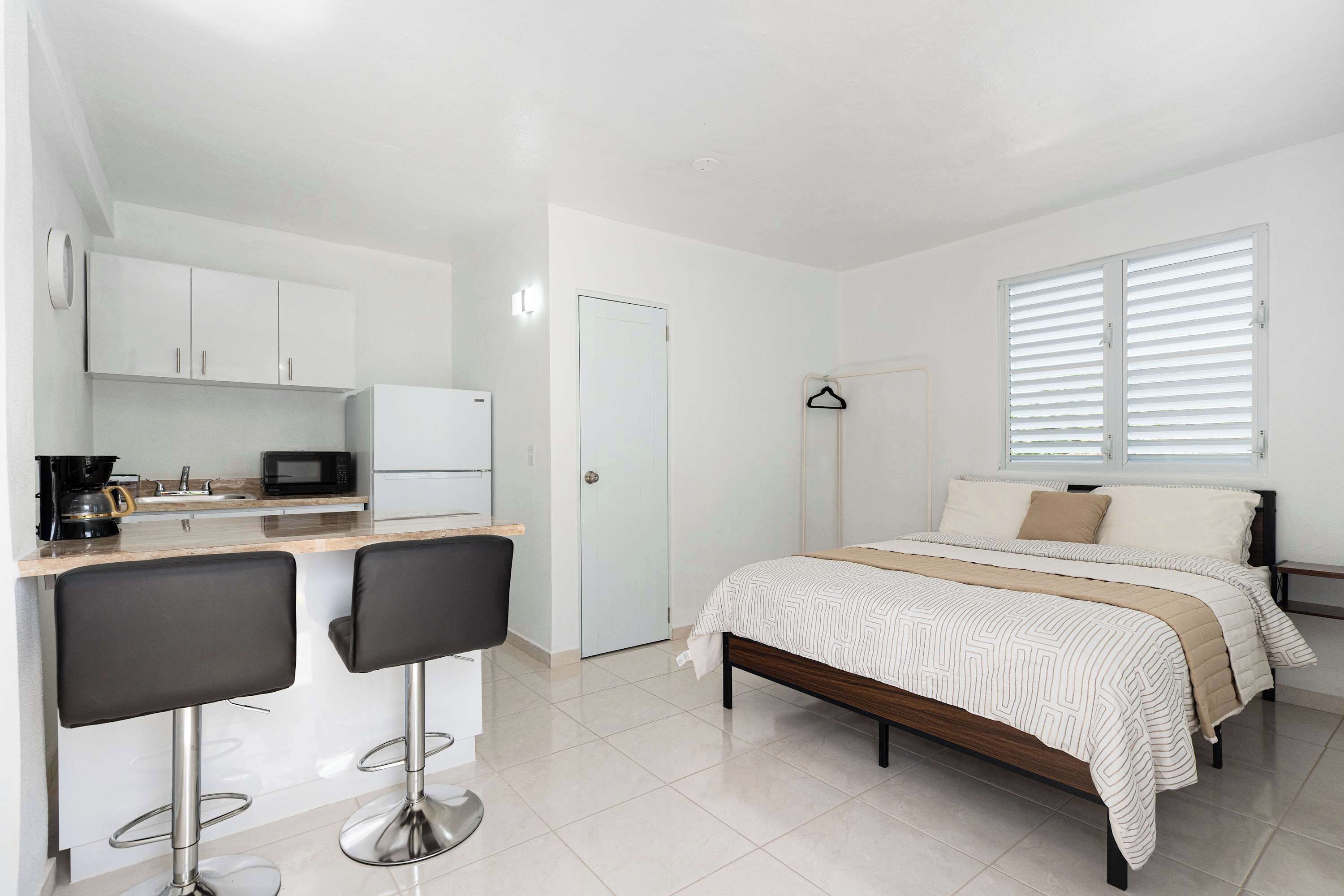 Un dormitorio pequeño con cama, mesitas de noche, una ventana con persianas y una kitchenette equipada con microondas y cafetera.