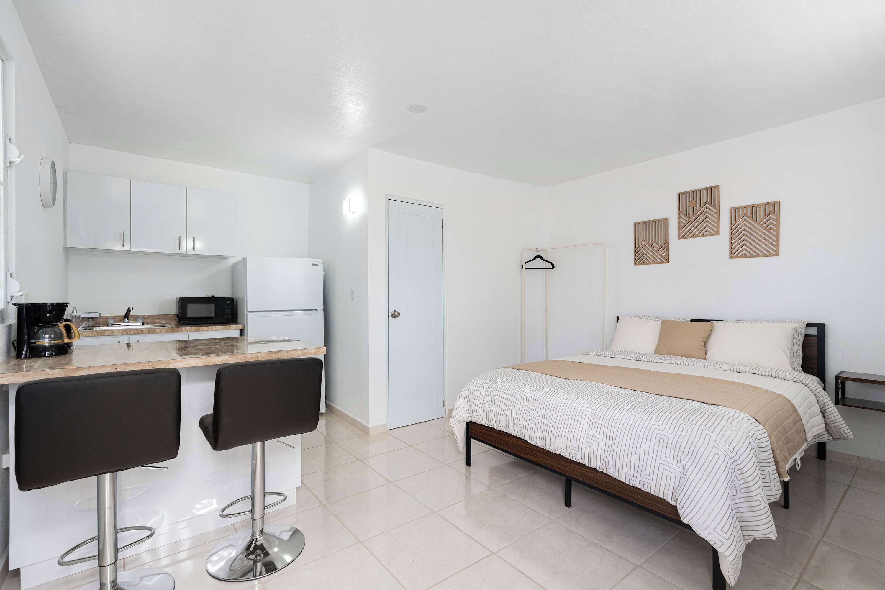Una habitación moderna y bien iluminada con kitchenette, una cama con colcha estampada y un perchero de pared.
