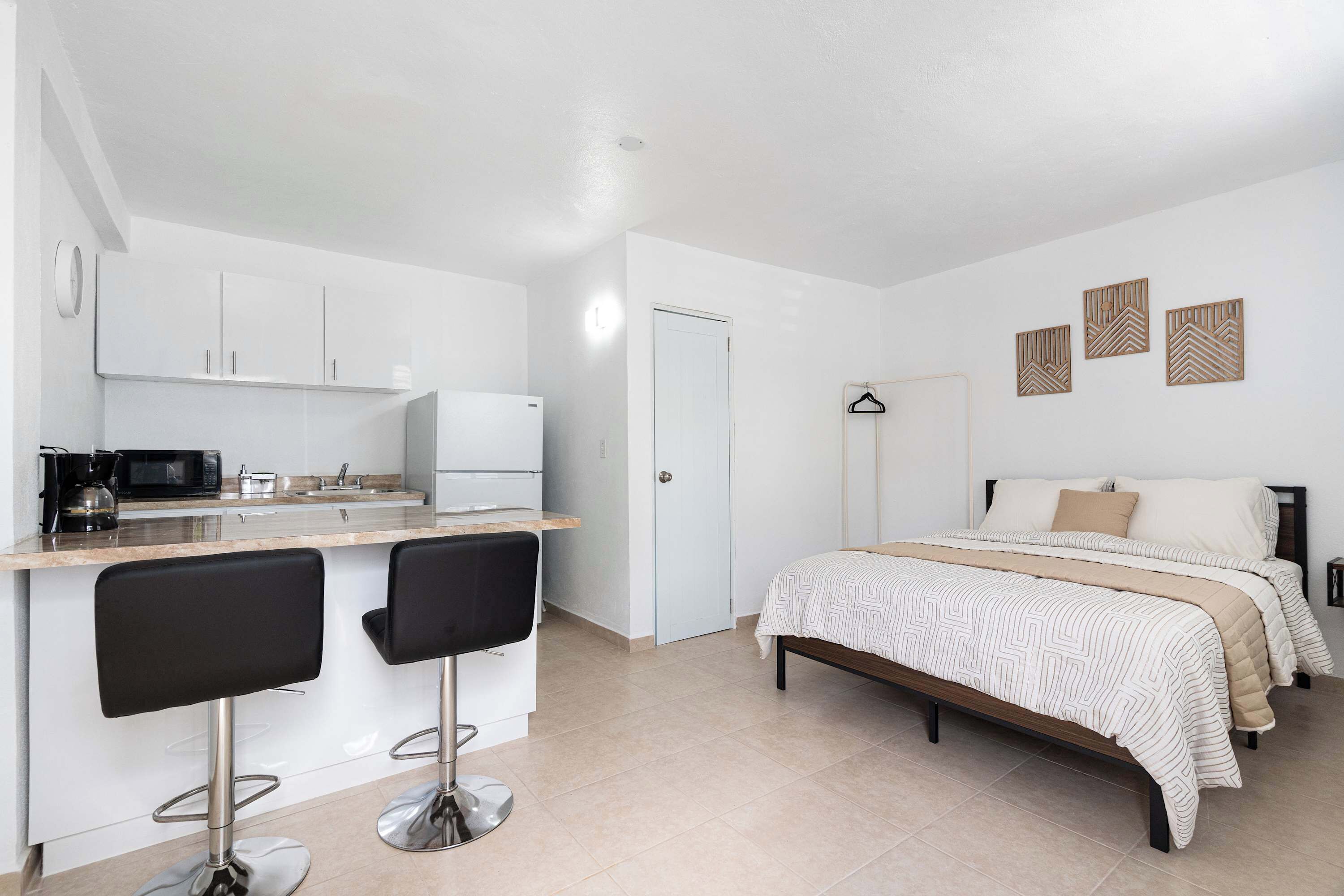 Una camera compatta con angolo cottura, un letto con copripiumino decorato e un piccolo angolo salotto con due poltrone.