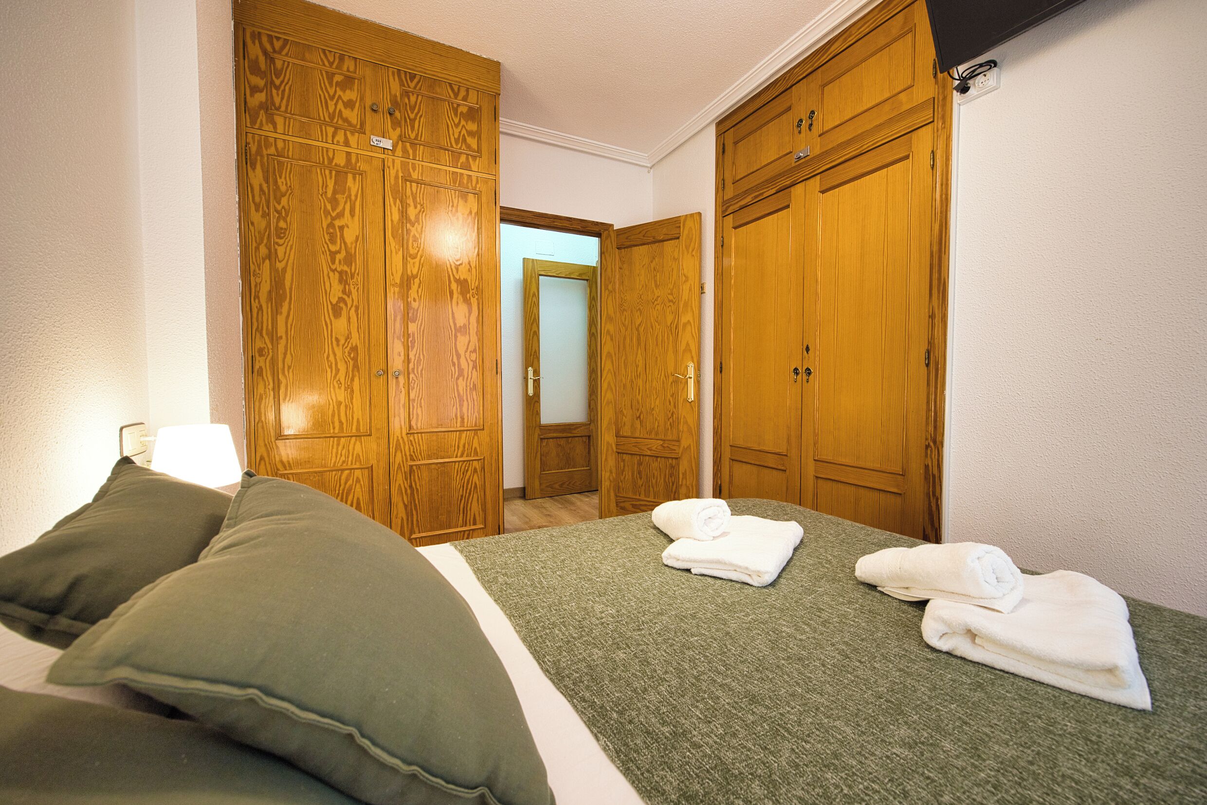 A hotel room with a bed, green bedding, two pillows, a pair of towels, and wooden wardrobes.