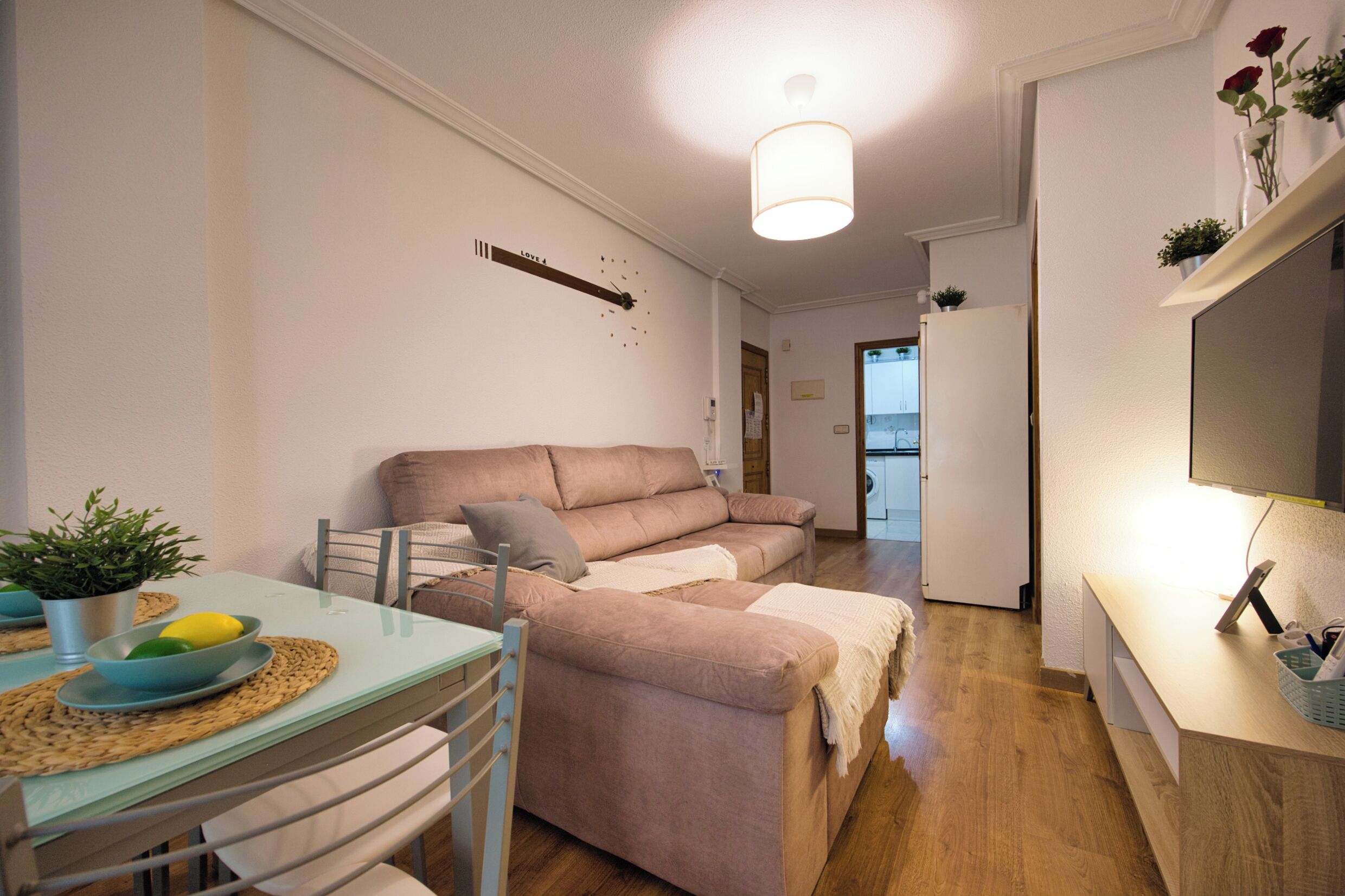 Een knus woongedeelte met een bank, een eettafel met stoelen, een kleine keukenhoek en een flatscreen televisie aan de muur.