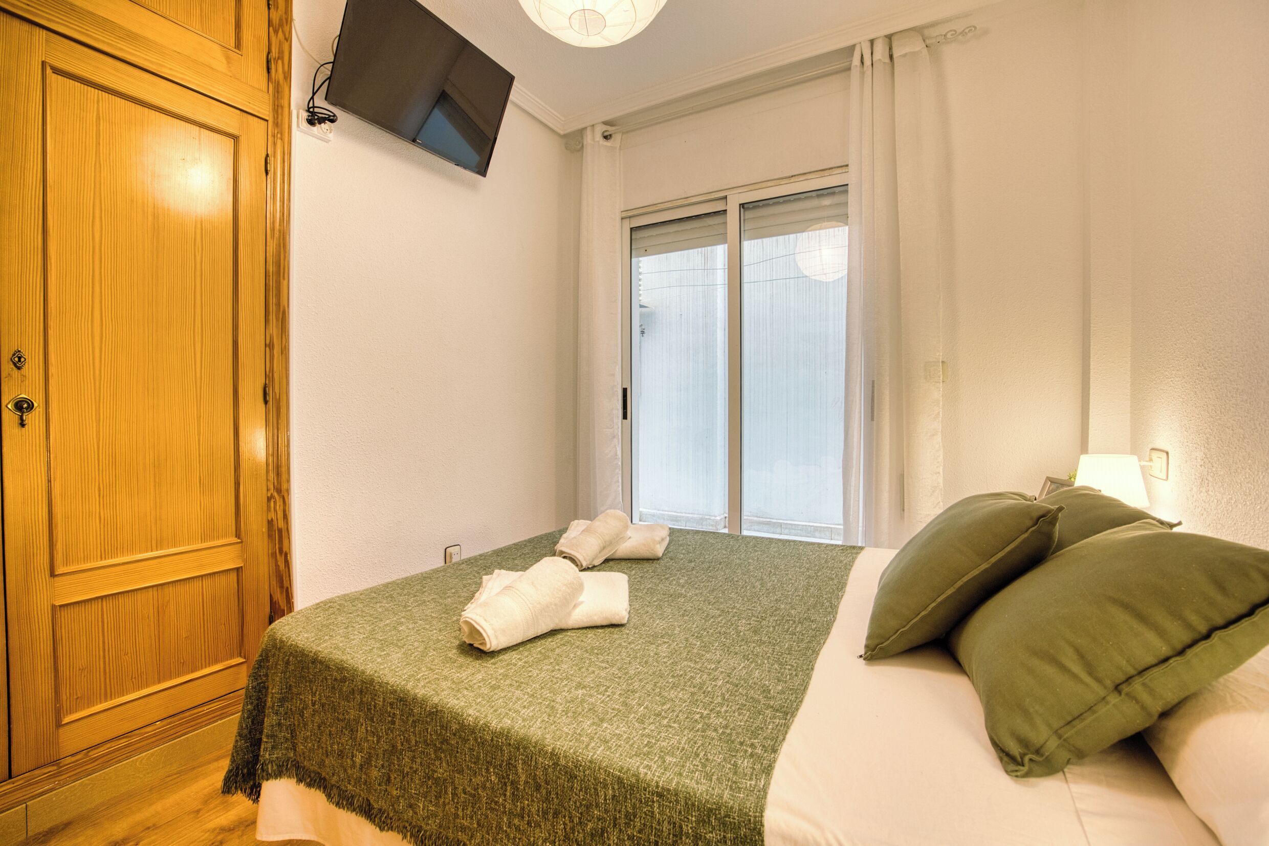 Ein Hotelzimmer mit Holzschrank, einem Bett mit grüner Bettwäsche, einem an der Wand montierten Fernseher und einem Fenster mit Vorhängen.
