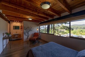 Ein Schlafzimmer mit großem Fenster, Holzbalken an der Decke, einem Bett, einem Sessel und einem kleinen Tisch mit Lampe.