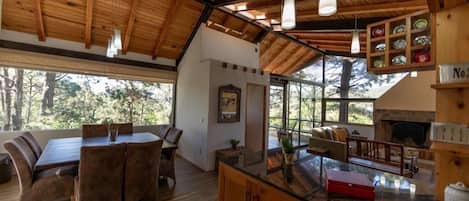 A spacious dining area with a wooden table and chairs, a kitchen with a stone fireplace, and a large window offering a view of the outdoors.
