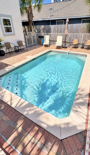 A rectangular swimming pool with a tiled edge, surrounded by a brick patio and a wooden fence.