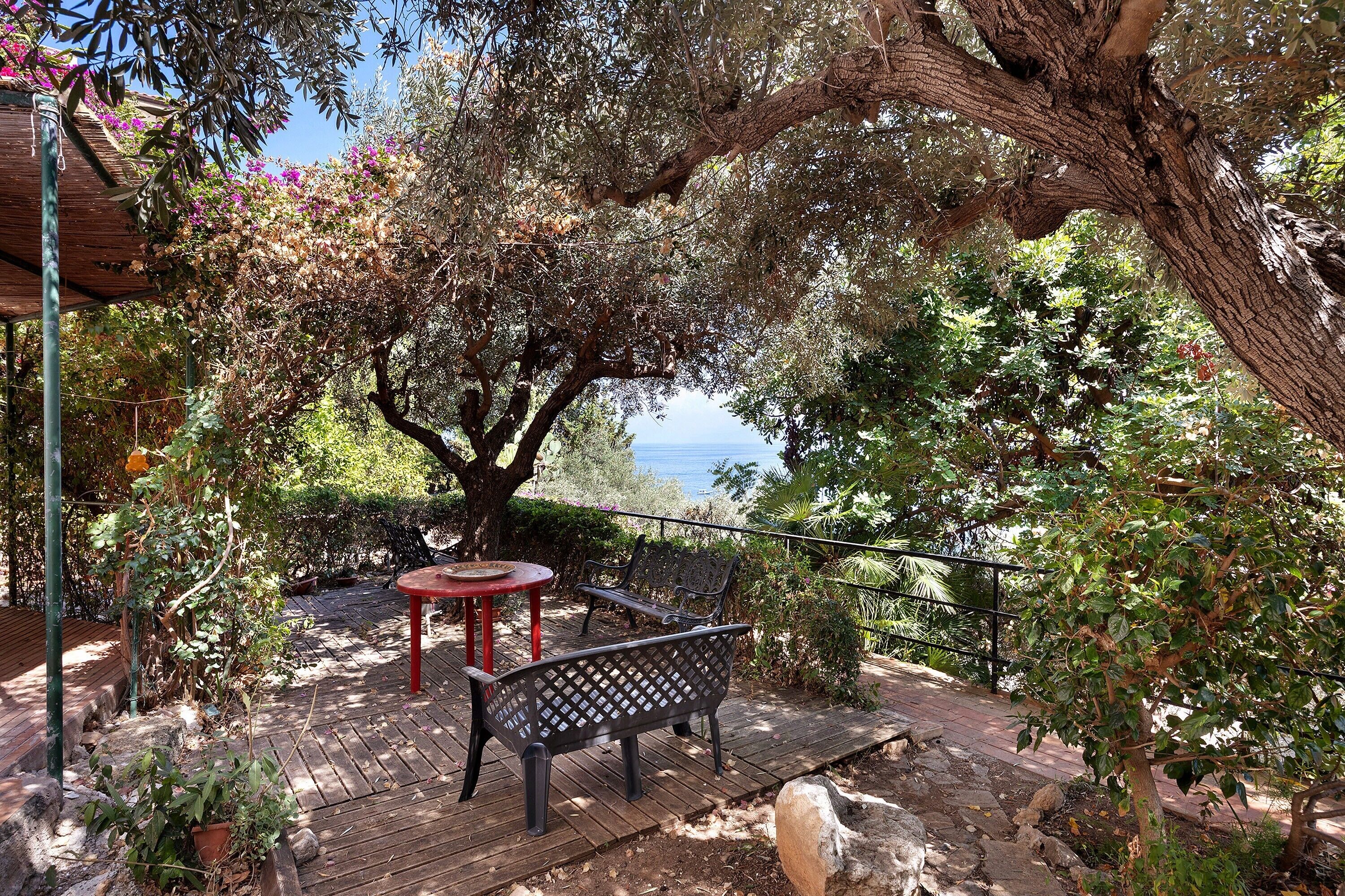 Et udendørs siddeområde med bænk og bord, omgivet af træer og udsigt til havet.