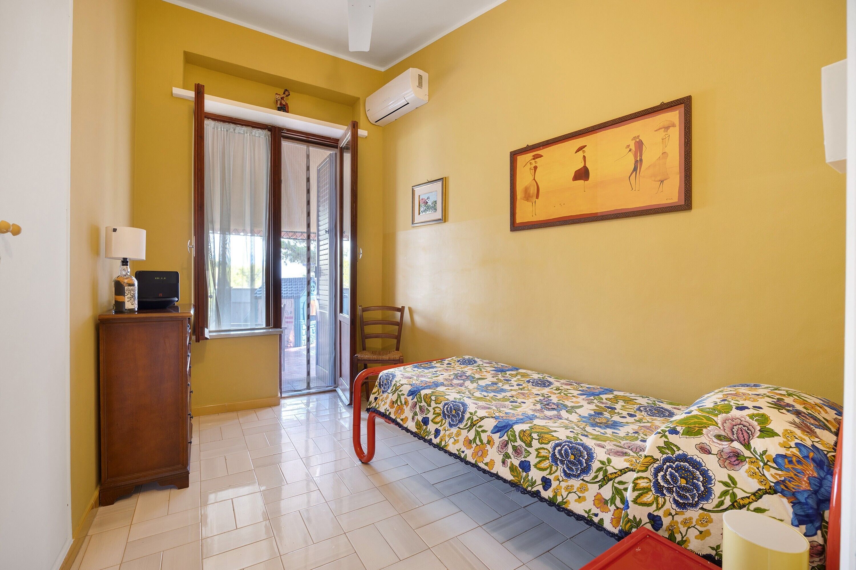 Una habitación con cama, una silla, una mesita, ventana con cortinas y aire acondicionado instalado en la pared.