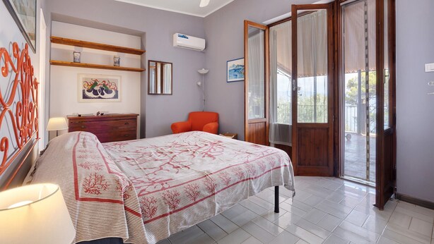 Une chambre à coucher comprenant un lit, une chaise, un bureau, une lampe, un ventilateur de plafond et une fenêtre avec des rideaux.