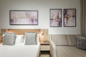 A hotel room with a bed, two pillows, a bedside table with a lamp, a sofa, and three abstract paintings on the wall. - One Rosebank: Where Style Meets Convenience (Johannesburg)